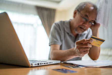 Depressed Asian Senior male feeling worried about financial problem. Stressed desperate elderly older man looking frustrated to paperwork and bills think of money debt, budget loss, bankruptcy at homeの写真素材