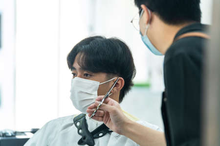 Asian hairdresser barber wear mask, cut hair of attractive man in shop. Handsome young man customer in modern salon barbershop, enjoy getting haircut service by stylist man during  pandemic.の写真素材