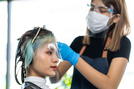 Asian beautiful girl changing new hair style and color in beauty salon. Professional Hairdresser barber wear face mask to prevent from pandemic dye woman customer hair for hairdo in barbershopの写真素材