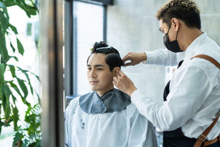 Asian hairdresser barber wear mask, cut hair of attractive man in shop. Handsome young man customer in modern salon barbershop, enjoy getting haircut service by stylist man during pandemic.の写真素材