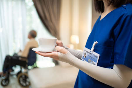 Asian beautiful therapist doctor serve coffee to old patient in house. Young woman nurse at nursing home taking care of disabled senior elderly old man on wheelchair. Medical insurance service conceptの写真素材