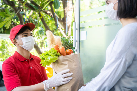Asian deliver man wear face mask in red uniform handling box of food, groceries, fruit give to woman customer in front of the house. Postman and express male grocery delivery service duringの写真素材