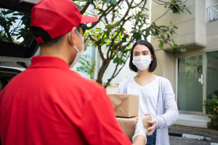 Asian postman, deliveryman wearing mask carry small box deliver to customer in front of door at home. Man wearing mask prevent , corana virus affection outbreak. Home delivery shopping concept.の写真素材
