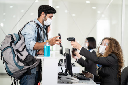 Male traveler shows phone to female officer at airline check in counter for issue airplane ticket boarding pass. Woman staff wear face mask to prevent from coronavirus pandemic. New normal lifestyleの写真素材