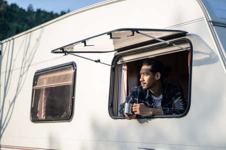 Asian young man feeling relax on camper van drinking coffee in the morning with happiness and smile on vacation. Male traveler looking outside the window to see the nature view, beautiful mountain.の写真素材