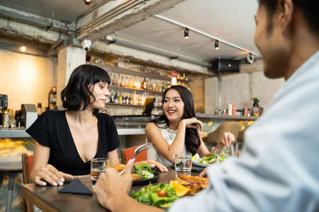 Group of young interracial best friend people sitting indoor restaurant eating food together for lunch. Women talking to Asian man about meal with smile. They are feeling happy and enjoying the meal.の写真素材