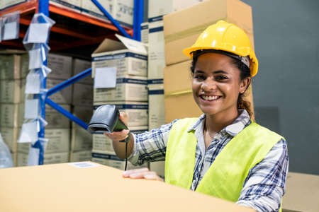 Asian female engineer worker working in shipping industrial. Young woman with safety hard hat, vest checking quantity of storage product on shelf in store warehouse factory. Girl works with smile faceの写真素材