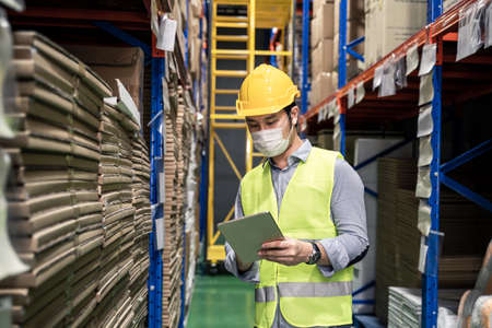 Asian male engineer worker wear mask work during pandemic in store warehouse shipping industrial. Young man with safety hard hat, vest checking quantity of storage product on shelf in factory.の写真素材