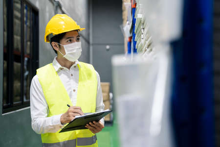 Asian male engineer worker wear mask work during pandemic in store warehouse shipping industrial. Young man with safety hard hat, vest checking quantity of storage product on shelf in factory.の写真素材