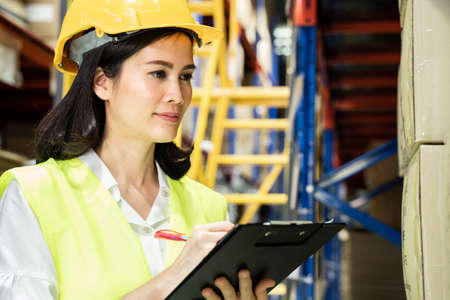 Asian female engineer worker working in shipping industrial. Young woman with safety hard hat, vest checking quantity of storage product on shelf in store warehouse factory. Girl wの写真素材