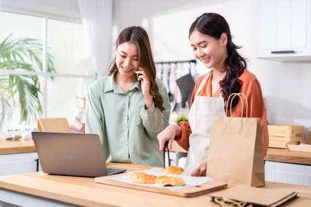Asian beautiful business woman and friend bake bakery and sale in house. Attractive lesbian couple feel happy and exciting for success in entrepreneur sell food order from home then high five togetherの写真素材