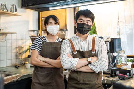Portrait of attractive Asian waiter and waitress at restaurant cafe. Barista business couple in apron wearing protective mask , stand with confident and crossing arm due to  at coffee shop.の写真素材
