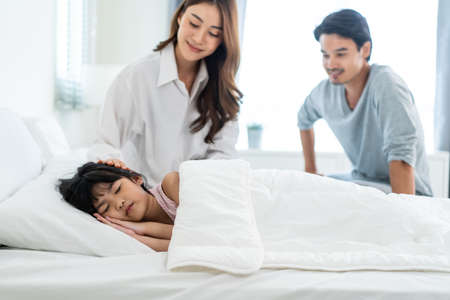 Asian loving parents take care of sleeping daughter in bedroom at home. Attractive happy family, beautiful couple put blanket on comfortable asleep napping little baby kid girl on bed in early morningの写真素材