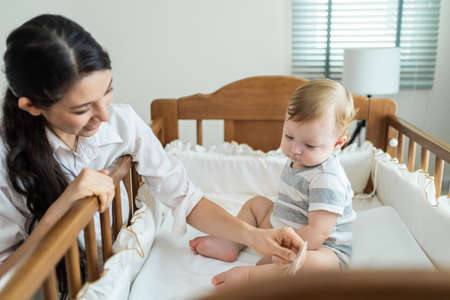 Caucasian mother play with cute baby boy child on infant bed at home. Happy family, attractive young woman mom talking to toddler son with happiness enjoy after wake up activity in bedroom in house.の写真素材
