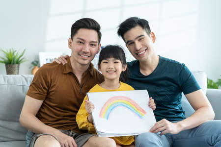 Portrait of handsome man gay family with young daughter in living room. Little adorable girl child sit on sofa with attractive romantic male lgbt couple and shows rainbow drawing picture to two fatherの写真素材