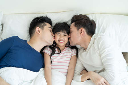 Portrait of handsome man gay family with young kid daughter in bedroom. Attractive romantic male lgbt couple lying down on bed and kiss little adorable girl child in morning, enjoy parenting in house.の写真素材