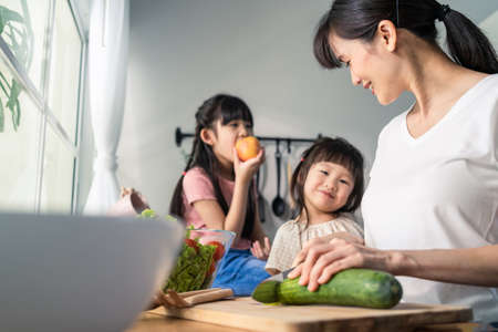 Asian beautiful mother cooking vegetable salad with two young daughter. Happy Family, attractive caring parent mom enjoy preparing healthy foods for little girl child sibling sister in kitchen at homeの写真素材