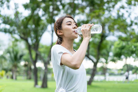 Asian young girl athlete drink bottle of water after exercise in park. Active attractive beautiful sportswoman take a break after yoga, running workout outdoors in evening for health care in garden.の写真素材