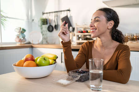 Latino attractive woman enjoy eating healthy foods in kitchen at home. Beautiful young female feel happy while sitting on table, eating fruits and clean water for health care and lose weight in house.の写真素材