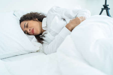 Latino beautiful sick girl in pajamas getting up from sleep in bedroom. Attractive young woman feel bad and painful after wake up on bed, having stomach ache or periods then clasping belly in morning.の写真素材