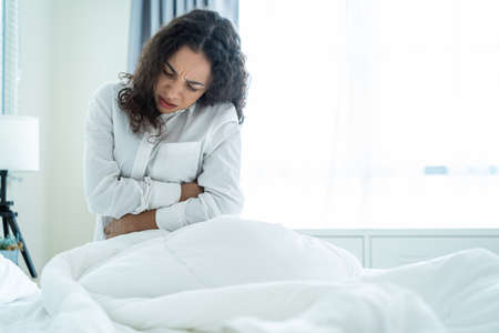 Latino beautiful sick girl in pajamas getting up from sleep in bedroom. Attractive young woman feel bad and painful after wake up on bed, having stomach ache or periods then clasping belly in morning.の写真素材