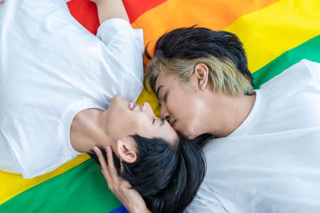 Asian man gay family lying down on LGBT flag and look at each other. Attractive handsome lgbt couple sleep on bed in bedroom in morning and kiss forehead of boyfriend with pride and rainbow backgroundの写真素材
