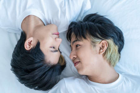 Asian happy man gay family lying down on bed and look at each other. Attractive handsome loving lgbtq couple sleep on bed in bedroom in early morning and kiss forehead of boyfriend with happiness.の写真素材