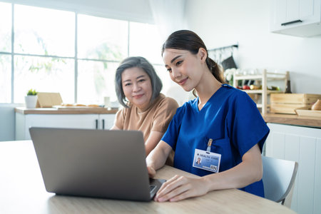 Asian nurse at nursing home take care of disabled senior elderly woman. Attractive beautiful therapist doctor use laptop computer and show diagnosis treatment plan to older grandma patient in house.の写真素材