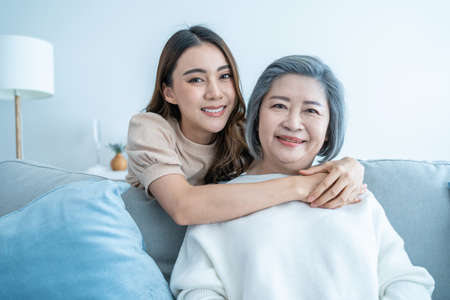 Portrait of Asian lovely family, young daughter hugging older mother. Attractive woman and senior elder mature mom sit on sofa, enjoy activity at home together in living room and smile, look at cameraの写真素材