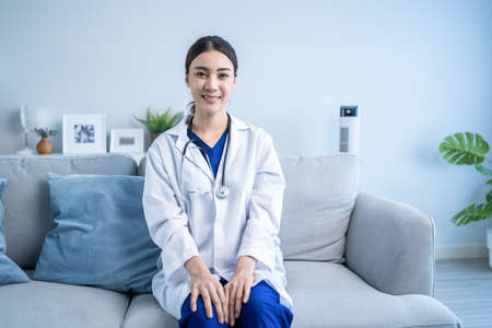Portrait of beautiful physician doctor wear gown sit on sofa in house. Attractive caregiver therapist practitioner pharmacist girl smile and looking at camera after success from treatment in hospital.の写真素材