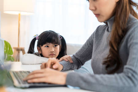 Asian young kid daughter feeling sad and need to play with busy mother. Attractive beautiful mom use laptop computer working from home during new normal and have no time for little child girl in houseの写真素材