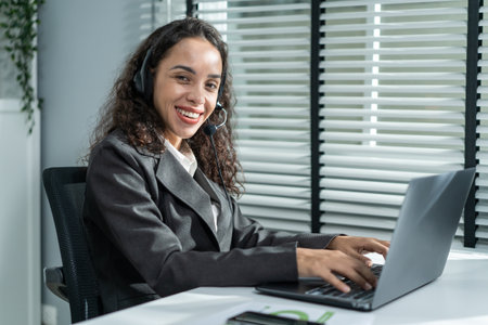Portrait of Latino beautiful business woman smile while work in office. Attractive professional female employee worker in formal wear sit on table at workplace, use laptop computer and look at camera.の写真素材