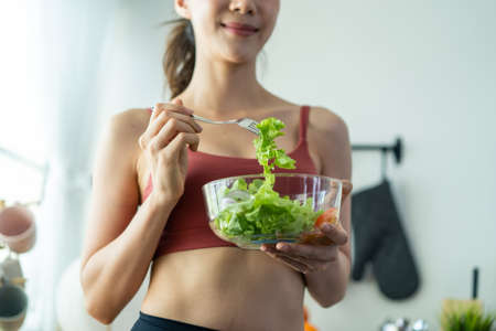 Close up of Asian attractive woman hold salad bowl and eat vegetable. Beautiful sport girl in sportswear enjoy eat clean vegetables after exercise for health in house. Diet and Healthy food concept.の写真素材