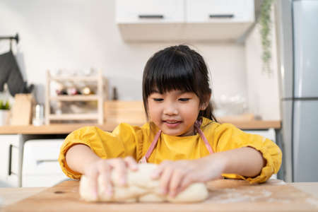 Close up Asian happy young kid girl doing homemade bakery in kitchen. Adorable little child sit on table feeling happy and enjoy learn to cooking foods or baking  kneads yeast dough with hands at homeの写真素材