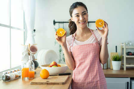 Portrait of Asian attractive woman holding orange and look at camera. Young beautiful girl wear apron, feel happy after cook fruit salad enjoy preparing healthy foods for breakfast in kitchen at home.の写真素材