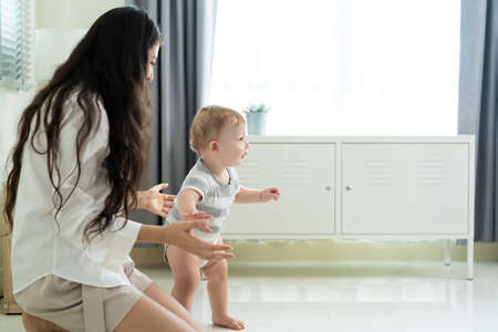 Caucasian cute little baby boy child learn to walk with mom in house. Happy family, mother support...