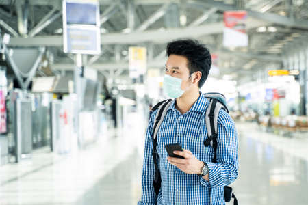 Asian traveler business man with luggage, bag wearing face mask, hold phone at departure check in airport terminal. Smart male passenger traveling by plane transportation during covid19 virus pandemicの写真素材