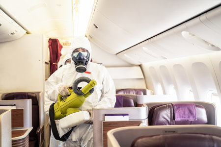 Cleaner officers hold cleaning device on airplane passenger cabin. An employee sprays disinfectant aboard a plane, during airline's sanitary measures to help curb the spread of pandemic.の写真素材