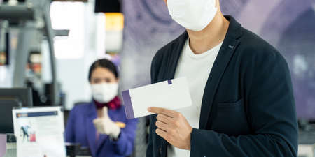 Asian male traveler hold boarding pass ticket at customer check at airline service counter. Man and woman wearing face mask when traveling by airplane transportation to prevent virus pandemic.の写真素材