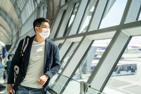 Asian traveler business man wearing face mask waiting to board into airplane, standing in departure terminal in airport. Male passenger traveling by plane transportation during covid19 virus pandemic.の写真素材