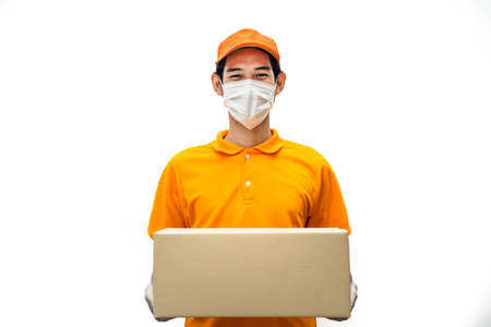 Asian deliver man wearing face mask in orange uniform holding box of food, groceries, things standing in white isolated background. Postman and express grocery delivery service during covid19 pandemicの写真素材