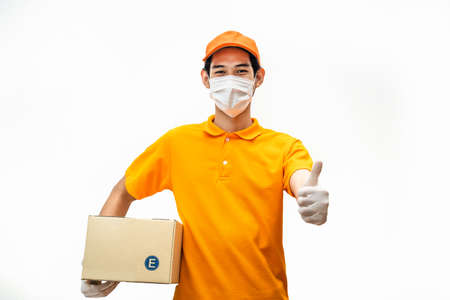 Asian deliver man wearing face mask in orange uniform holding box of food, groceries gives thumbs up standing in white isolated background. Postman and express delivery service during pandemicの写真素材
