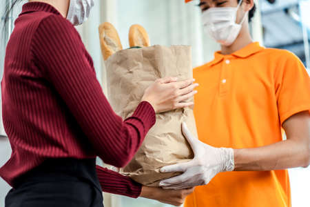 Asian deliver man wearing face mask in orange uniform handling bag of food, groceries, fruit give to woman costumer in front of the house. Postman and express grocery delivery service during covid19.の写真素材
