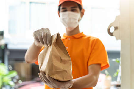 Asian deliver man wearing face mask in orange uniform holding bag of food, groceries, fruit standing in front of customer home. Postman and express grocery delivery service during covid19 pandemic.の写真素材