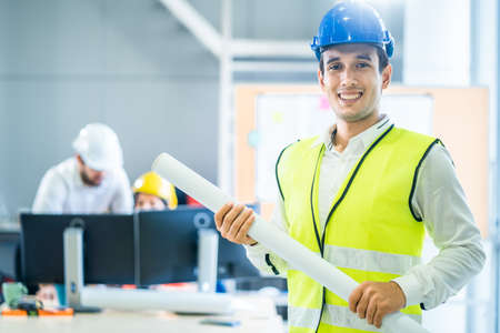 Asian man hold blueprint drawing while his team members looking project area. Young engineer group of home building member discuss for project planning.People brainstorm teamwork for idea of structureの写真素材