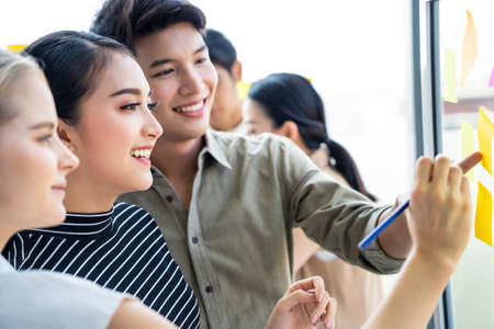 Group of diversity people brainstorm share knowledge in team. Asian beautiful woman giving idea with smile, female colleague writing on sticky note on board. Young man friend enjoy working as teamworkの写真素材