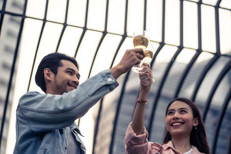 Asian young couple having celebrate anniversary party together on rooftop. Man and woman holding glass of white wine toasting and drinking with happiness and smile. Love of young boy and girl concept.の写真素材
