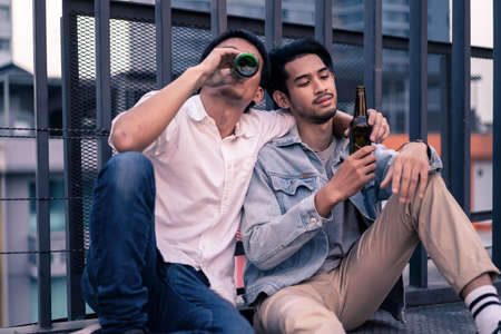 Couple men friends drinking alcohol beer on rooftop. Asian man having problem and trouble drunk and feeling bad crying and be consoled by his friend sitting next to him. Alcohol drの写真素材