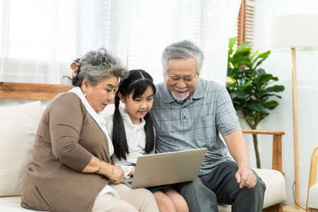 Asian grandparent with little young cute grandchild sitting on sofa playing laptop together. Little granddaughter teach senior elder to surf internet using computer and technology& modern lifestyle.の写真素材