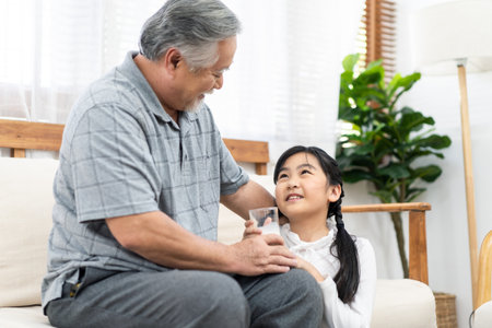 Asian little granddaughter bringing glass of milk to grandfather. Young niece girl take care of grandpa with happiness and love with smile. Elder health care and senior family retirement lifestyle.の写真素材